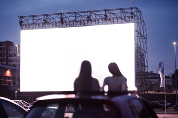 Rear view of two female friends sitting in the car while watching a movie in an open air cinema...