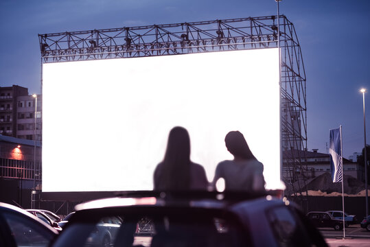 Rear view of two female friends sitting in the car while watching a movie in an open air cinema with a big white screen
