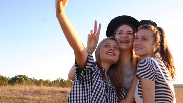 Three cheerful young women take pictures of their front camera on a field at sunset. Cheerful girls take selfies and smile in front of the camera.