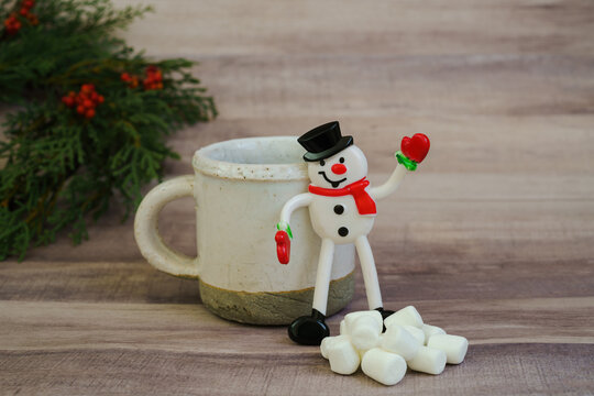 A snowman toy waves hello near a mug of cocoa and a pile of marshmallows in a Christmas setting that allows for copy space.