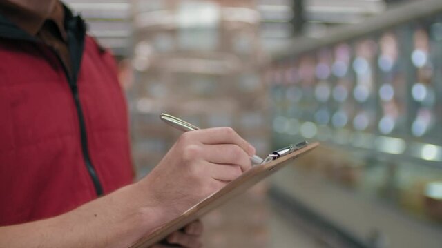 Midsection close-up of unrecognizable male merchandiser in hypermarket uniform analyzing stock plan making notes on paper