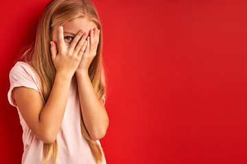 frightened girl in studio. she close her face. caucasian child stand afraid of something, looks at...
