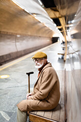 senior man in coat and cap looking away while sitting on underground platform bench