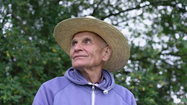 Mature farmer in strawy hat with big brim stands outdoor in farmland and looks arround