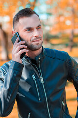 Handsome Young Man Talking on the Phone at the Beautiful Autumn Park. Youthful Guy Using Smartphone for the Call Outdoors at Sunny Day