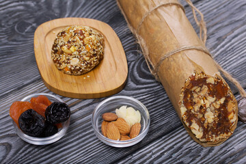 An energy bar made of dried fruits, nuts and seeds. Nearby there are ingredients for cooking, dried apricots, almonds and prunes. On black pine boards.
