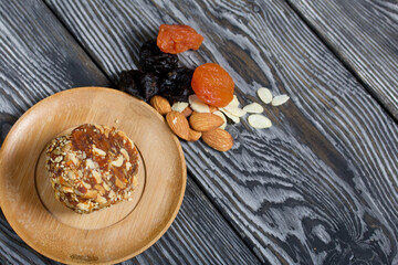 An energy bar made of dried fruits, nuts and seeds. Nearby there are ingredients for cooking, dried apricots, almonds and prunes. On black pine boards.