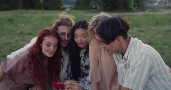 Happy millennial people using smartphone frontal camera while sitting at grass. Diverse good looking friends smiling while taking selfie and looking at photo. Concept of friendship