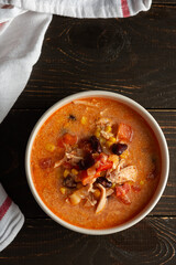 Spicy mexican soup with beans and corn, tomatoes and chicken breast, bowl of soup and napkin on dark wooden background, top view