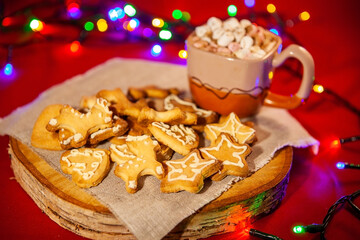 Christmas gingerbread cookies on a wooden stand and a cup of hot chocolate with marshmallows in the back, on a red background with a beautiful bokeh of glowing garland. Christmas and New Year concept.