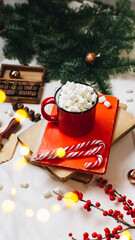A red mug with cocoa and marshmallows stands on a stack of old books. Nearby are spruce branches, New Year's decorations, a branch of ikles and stamps for letters. Christmas mood and a hot drink.