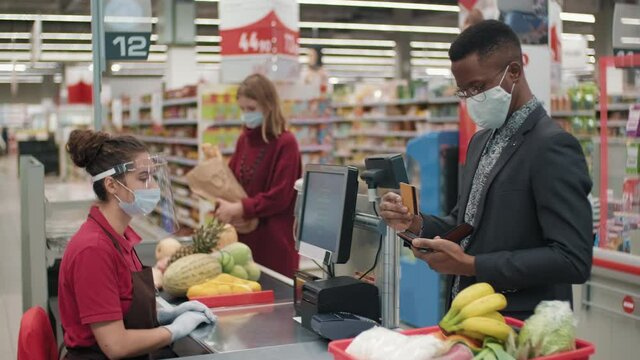 Medium shot of businesslike african american man in protective mask paying for food products contactlessly with credit card standing at cash desk in big supermarket