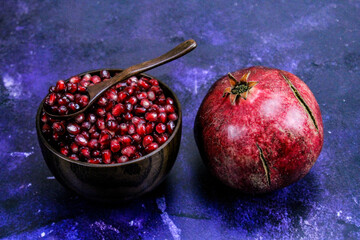 A bowl full of pomegranate seeds with a wooden spoon on it. A whole ripe pomegranate next to the...