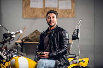 A young biker in a leather jacket in a garage smiles for a photo and his blog