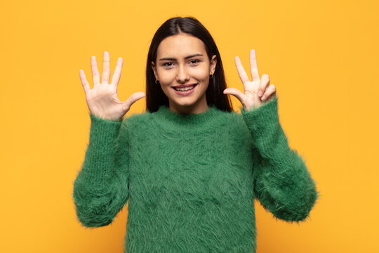 Young Hispanic Woman Smiling And Looking Friendly, Showing Number Eight Or Eighth With Hand Forward, Counting Down