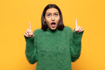 young hispanic woman looking shocked, amazed and open mouthed, pointing upwards with both hands to...