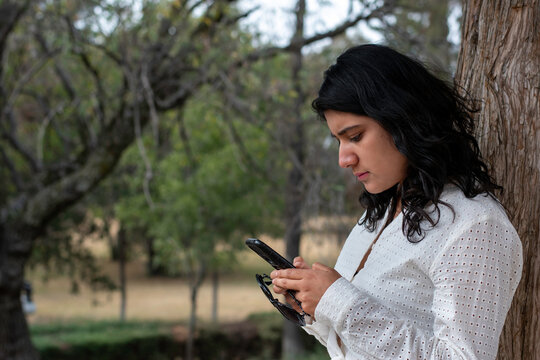 Mujer joven con vestido blanco apoyada en un árbol revisando su teléfono celular