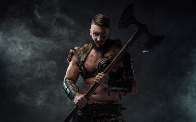 Fototapeta premium Northern vandal seafarer with muscular build holding huge axe and staring at camera in dark smokey background.