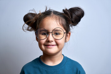 Fototapeta premium Portrait of cute smiling swarthy caucasian little girl with glasses and funny hairstyle, beautiful happy child looking at camera isolated in studio close up