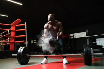 An African American man sprays magnesium or talcum powder with his palms before lifting a barbell....