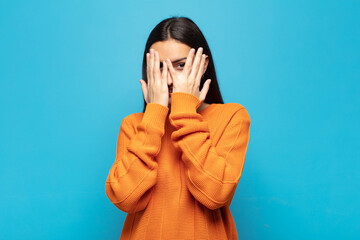 young hispanic woman covering face with hands, peeking between fingers with surprised expression...