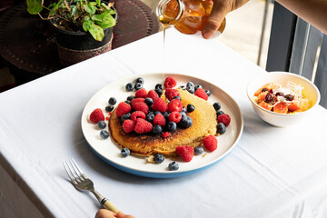 Breakfast at home, pouring honey into a vegan hot cake with raspberries and blueberries during the morning