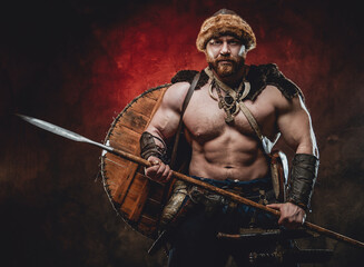 Fototapeta na wymiar Nude handsome northern barbarian with naked torso and muscular build poses holding a spear in dark red background.