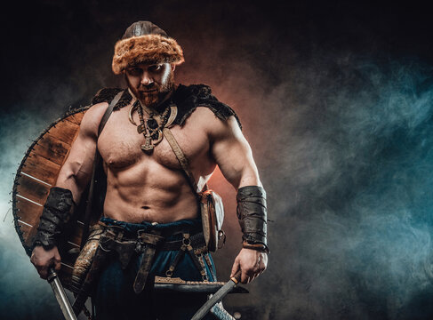Mad and savage northern warrior with naked torso and shield on his back poses in foggy background staring at camera and holding hatchet and knife.