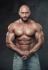 Fototapeta na wymiar Muscular and bald caucasian man with perfect abs and huge biceps poses in dark and smokey background looking at camera with serious face.