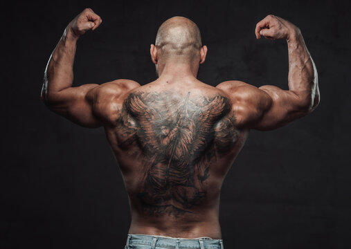 Shirtless and powerful bodybuilder poses in dark background with raised hands showing his muscular and tattooed back.