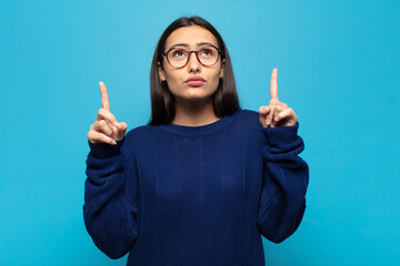 young hispanic woman looking shocked, amazed and open mouthed, pointing upwards with both hands to...