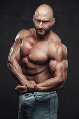 Fototapeta na wymiar Bearded and hairless man with powerful and muscular build in jeans shorts poses with angry face in dark background looking at camera.