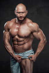Fototapeta na wymiar Caucasian muscular man with hairless head and naked torso pulls his shorts showing his muscular leg looking at camera with serious face in dark background.
