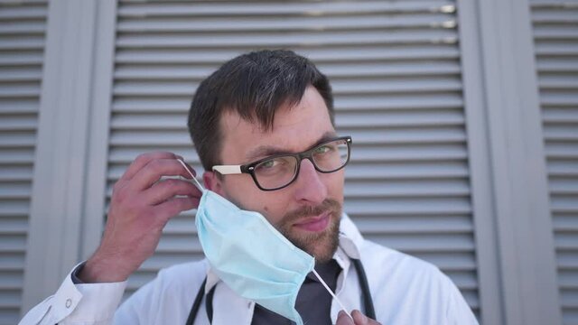 Young caucasian tired doctor putting on protective medical mask on his face, outdoor after coffee break before going to work in hospital. Serious, overworked, health care worker put in on mask