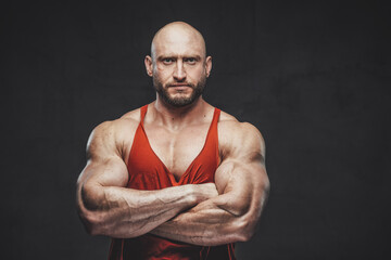 Powerful and hairless guy in red shirt with beard and strong hands poses in dark background looking...