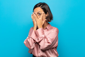 young hispanic woman covering face with hands, peeking between fingers with surprised expression...