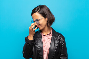 young hispanic woman feeling disgusted, holding nose to avoid smelling a foul and unpleasant stench