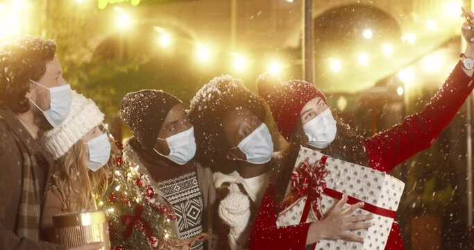 Cheerful multinational company of friends in medical masks standing in city center on winter evening taking joint photo holding gifts in their hands.