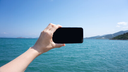 Horizontal smartphone. Tourist hand holding a black blank space screen smart phone while in vacation in the beach with view of the water blue landscape. Coast of Turkey, Aegean Sea