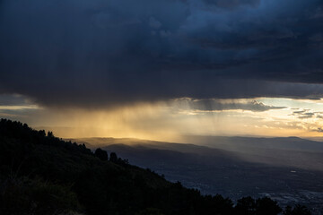 From the top of a mountain, there is a wide view during sunset time (golden hour). The gray clouds full of rain collide with the sunlight creating suggestive contrasts with plays of light.