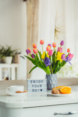 Good morning concept. Romantic breakfast - fresh flowers, cup of hot drink, cookies, orange, lightbox with message Enjoy your day on marble table with light interior view. Vertical card. Copy space.