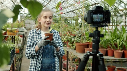 Slow motion of cute girl vlogger holding plant recording video in greenhouse waving hand showing like gesture using camera. People and vlogging concept.