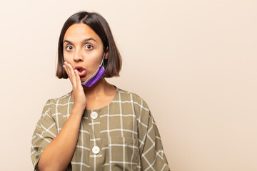 young hispanic woman feeling shocked and astonished holding face to hand in disbelief with mouth...