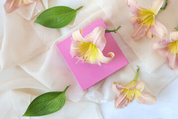 Pastel daylily flowers with a gift in a pink package, white background, top view, space for text-the concept of pleasant greetings on holidays