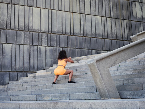 Young girl with curly hair doing squats on stairs. Outdoor sport. Orange sportswear.