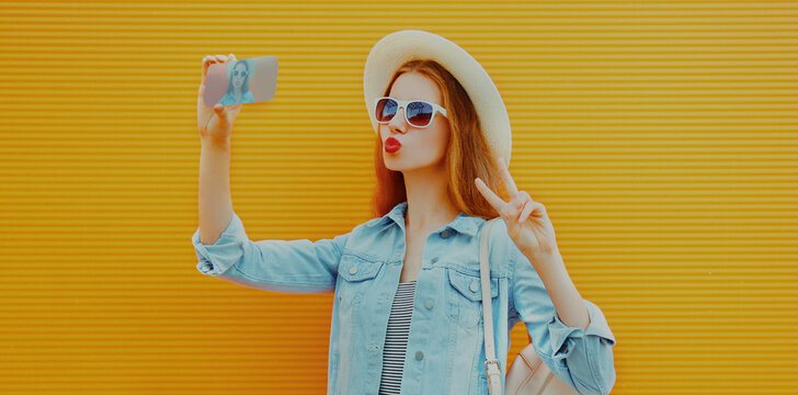 Portrait of young woman taking selfie picture by phone wearing a straw hat in the city over an orange background