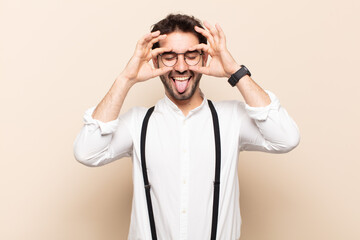 young handsome man smiling happily with funny face, joking and looking through peephole, spying on...