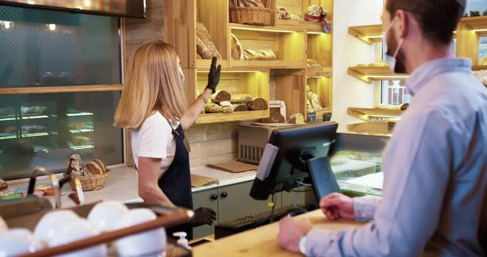 Caucasian female bakery shop worker in medical mask stands at work and speaking with male customer selling bread. Small business concept. Woman in apron at the counter in bakery shop. Food concept