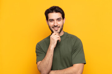 young handsome man looking happy and smiling with hand on chin, wondering or asking a question,...