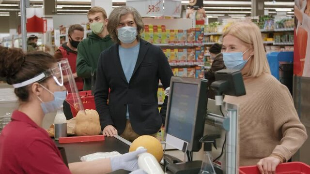 Medium footage of customers with shopping carts and baskets standing in queue wearing disposable masks while young female cashier scanning products in hypermarket